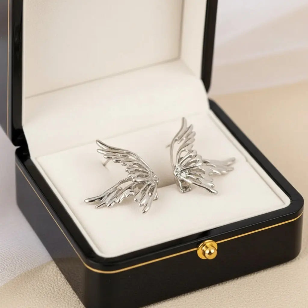 Silver wing-shaped earrings on a white surface with a soft pink fabric background